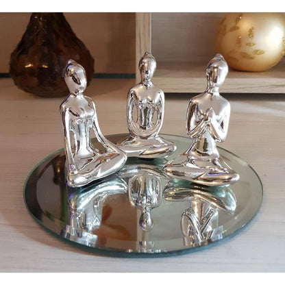 Três esculturas femininas prateadas em posições de meditação sobre espelho redondo