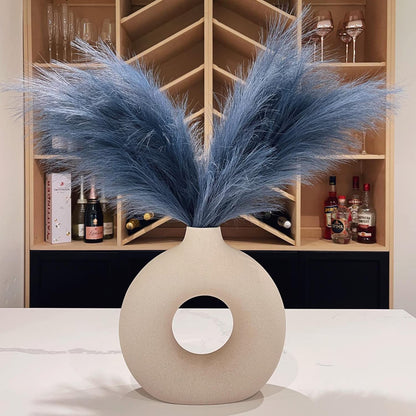 Vaso decorativo bege com abertura circular cheio de pampas azul em mesa branca em ambiente elegante