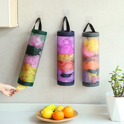 Dispenser de sacolas plásticas coloridas pendurado na parede da cozinha com mão puxando uma sacola, frutas e planta