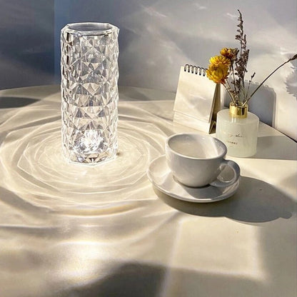 Luminária de vidro transparente com luz LED em mesa branca ao lado de xícara de café e vaso com flores secas