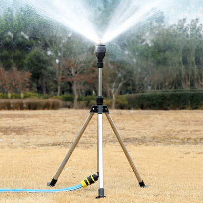 Aspersor de jardim de metal com mangueira azul irrigando grama seca e árvores ao fundo